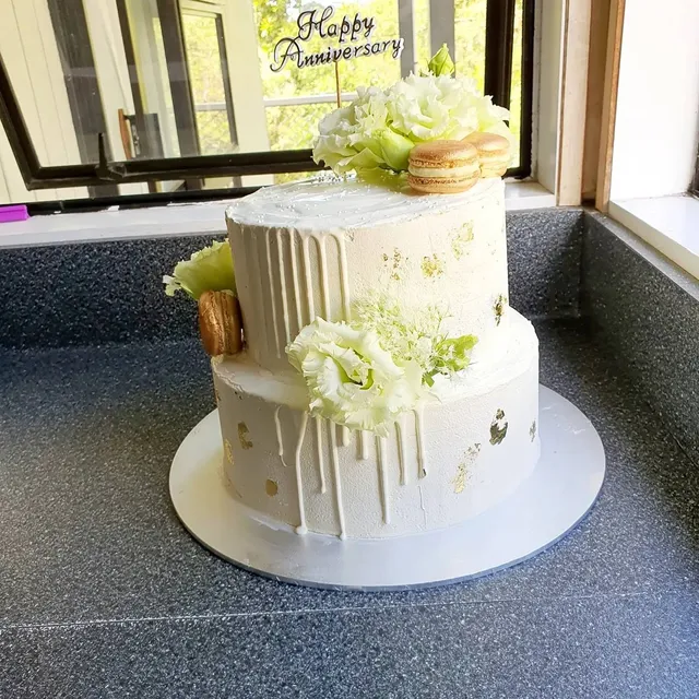 Two-tier white buttercream anniversary cake with gold leaf, macarons, and fresh blooms